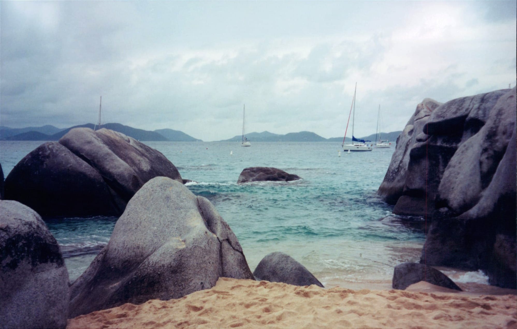 Giant boulders on a beach with sailboats beyond