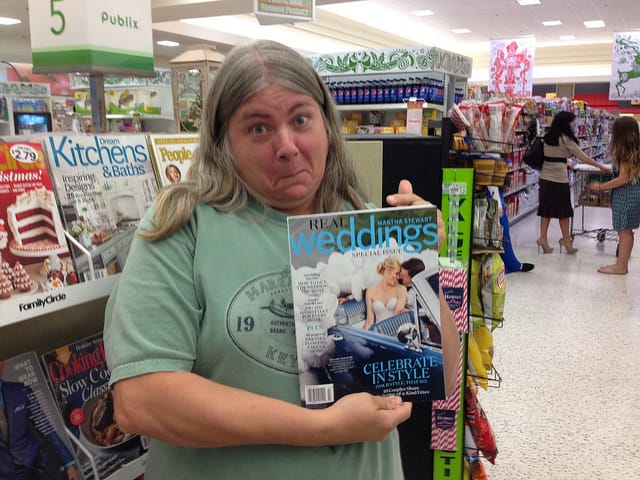 Woman with incredulous expression holding up a Real Weddings magazine at a grocery checkout