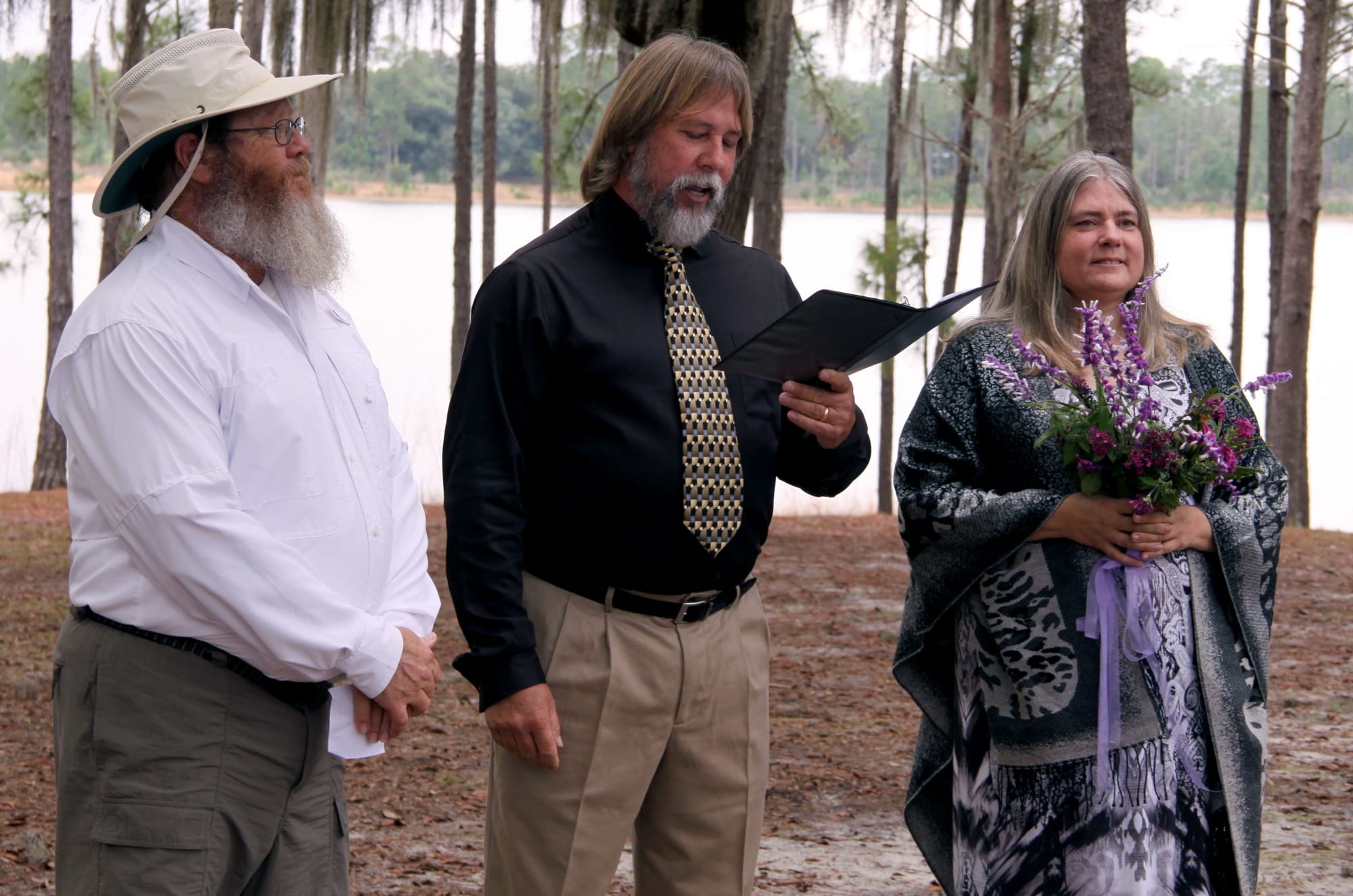 Wedding ceremony with groom, officiant, and bride among trees by a lake