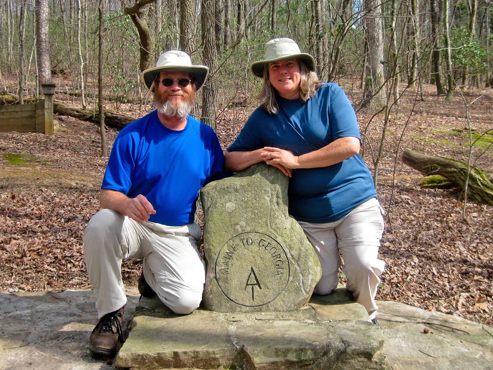 A man and a woman in hiking clothes posing with a stone monument that says Maine to Georgia AT
